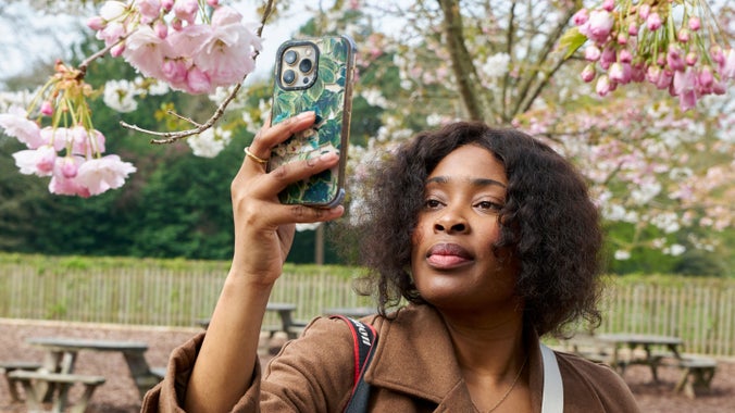A visitor holds up her phone to photograph a cluster of pink blossom on the tree she stands under.